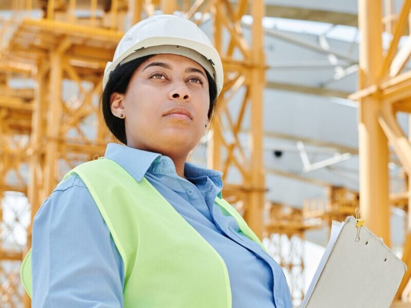 Confident female engineer in safety gear at a busy construction site, holding a clipboard.